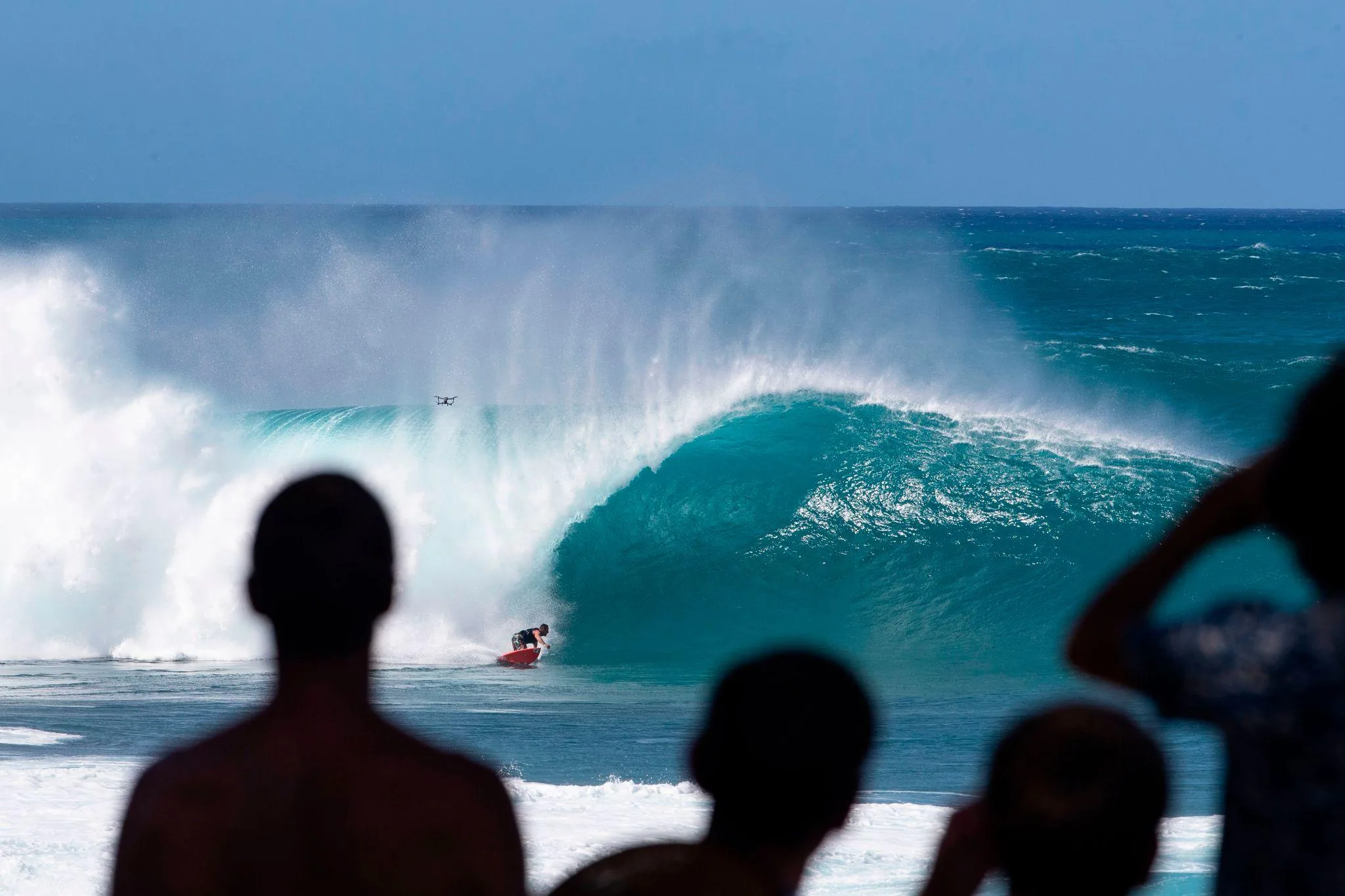 A large winter wave curling and breaking on the North Shore as a surfer rides it, watched by spectators silhouetted on the shore