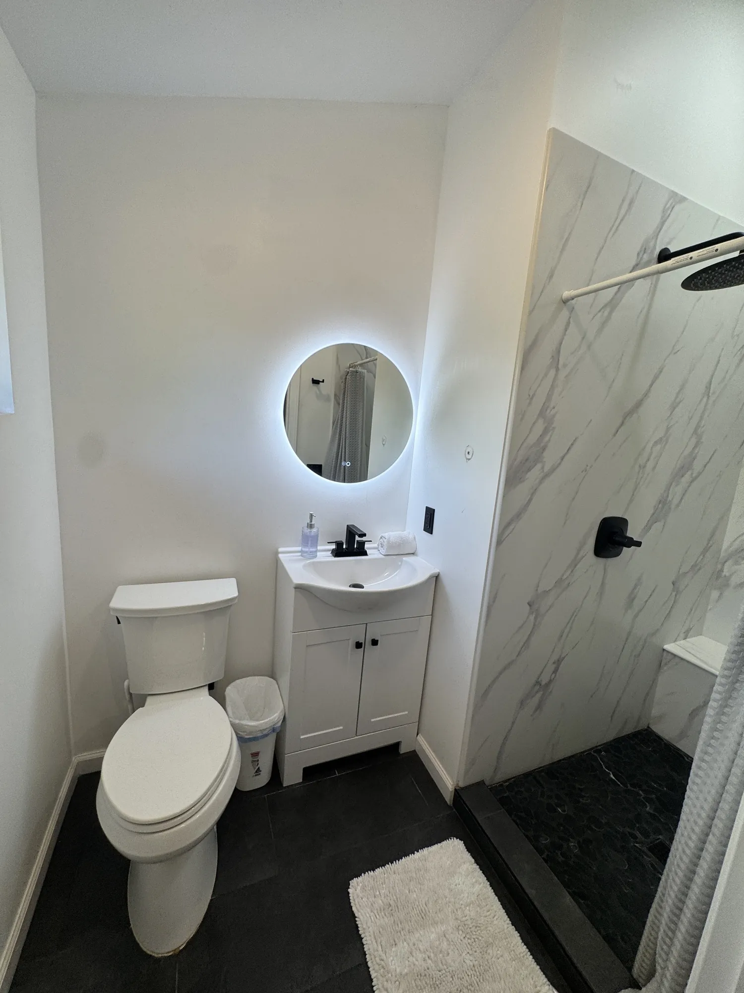 A bathroom with a round backlit mirror, vanity and marble-tiled walk-in shower