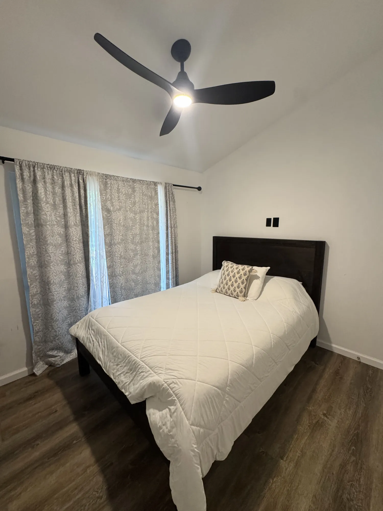 A bedroom with a dark wood bed frame, ceiling fan and curtained window