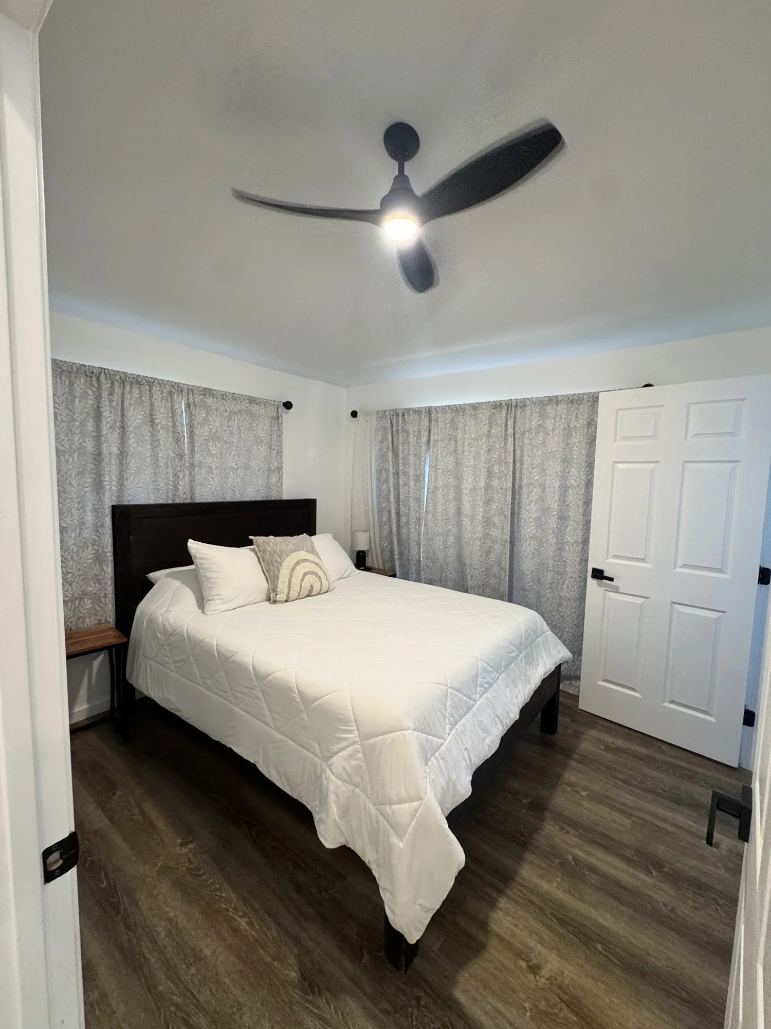 A bedroom with a ceiling fan, curtained window and doorway to the hall